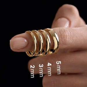 Könnte beinhalten: Vier glatte, polierte goldfarbene Ringe, die auf einem Finger gestapelt sind und mit Breiten von 2 mm, 3 mm, 4 mm und 5 mm beschriftet sind. Die Ringe haben ein abgerundetes, gewölbtes Profil und eine reflektierende metallische Oberfläche.