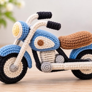 Könnte beinhalten: Ein gehäkeltes Motorrad-Spielzeug mit blauem Rahmen, schwarzen Reifen mit weißen Zentren, einem braunen Sitz und cremefarbenem Lenker. Das Design umfasst einen gelben Scheinwerfer, ein kleines rotes Rücklicht und einen weißen Motorblock.