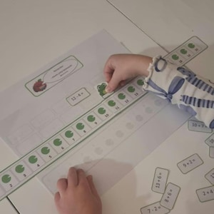Puede incluir: Una hoja de actividades de matemáticas con una recta numérica con iconos de nenúfares verdes numerados del 2 al 20. Un pequeño recorte de rana está colocado en la recta. Hay tarjetas de ecuaciones dispersas.