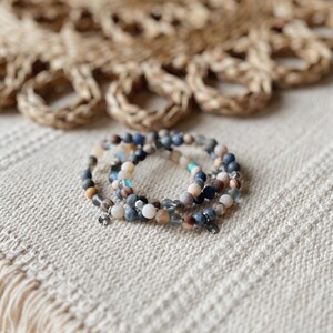 May include: Three beaded bracelets stacked on top of each other. The bracelets are made of blue, brown, white, and grey beads. The bracelets have silver charms.