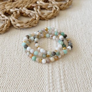 May include: Three beaded bracelets featuring a mix of white, pale blue, and brown beads, with gold-coloured accents. The bracelets are stacked on a white textured surface, with a woven straw element in the background.