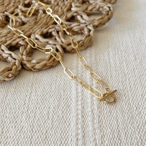 May include: A gold-coloured chain necklace with an oval link design and a toggle clasp. The necklace is displayed on a textured, off-white surface with a woven, tan background.