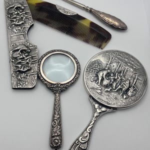 May include: An assortment of antique silver vanity items, including a comb, nail tool, hand mirror, and magnifying glass. The comb is tortoiseshell, and the mirror features a detailed relief scene. The items are arranged on a light background.