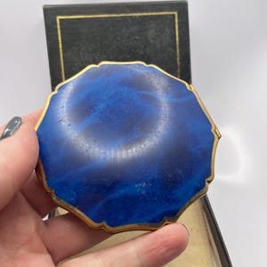 May include: A vintage compact mirror with a deep blue, marbled design. The octagonal compact has a gold-toned trim and is set against a black box with gold accents. The compact is held in a hand.