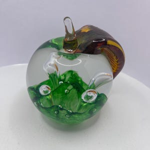 May include: A decorative glass apple sculpture. The apple has a clear, frosted body with green and white accents, and a brown and yellow swirl on top. The stem is clear glass.
