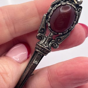 May include: A vintage silver brooch featuring a deep red oval gemstone. The brooch has intricate scrollwork and a long pin. The gemstone is approximately 2 cm in length. A classic piece of jewelry.