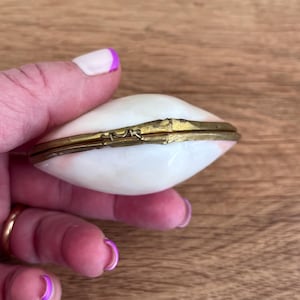 May include: A small, oval-shaped trinket box with a white and pink marbled design. The box has a gold-colored metal trim and a hinged lid. The box is held in a hand with painted nails.