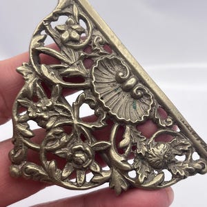 Op de afbeelding: Een messingkleurig, driehoekig decoratief metalen stuk met ingewikkelde bloemen- en bladerpatronen. Het open ontwerp toont rozen, gestileerde bloemen en bladeren. Het stuk heeft een licht verouderd uiterlijk.