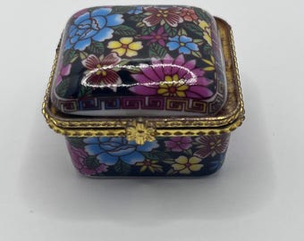 May include: A small, square trinket box with a floral design. The lid and sides are adorned with colorful flowers in shades of blue, pink, and yellow against a dark background. The box has a gold-colored metal trim.