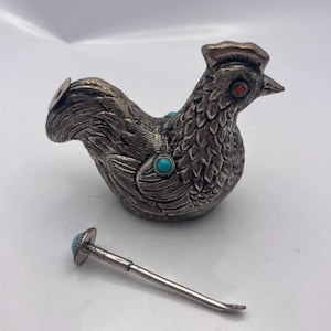 May include: A silver-toned rooster-shaped container with turquoise and red accents. The container has a detailed feather design and a small, matching applicator. The rooster's eye is a small red stone.