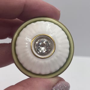 May include: A round, white ceramic cabinet knob with a central, sparkling, clear gemstone. The knob has a gold-colored ring around the gemstone and a green and gold border. The knob is held by a person.