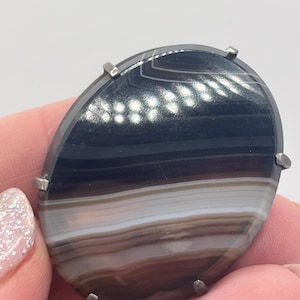 May include: Oval agate brooch with a silver-tone metal frame. The stone displays bands of black, brown, white, and tan. The brooch is held in a hand with light-coloured nail polish.