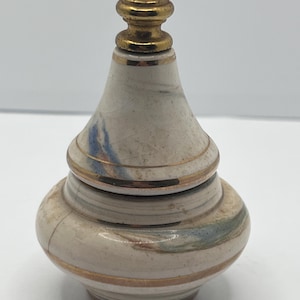May include: A small, decorative ceramic jar with a lid. The jar is off-white with blue and brown marbling and gold-colored bands. The lid is topped with a gold-colored finial. The jar has a bulbous base and a tapered body.