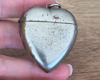 May include: A heart-shaped silver-toned metal locket or small container with a hinged lid. The surface shows visible signs of wear, including tarnishing and patches of discoloration, with a small metal loop at the top for attachment to a chain.
