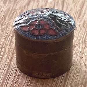 Può includere: Un piccolo contenitore cilindrico in metallo con un coperchio caratterizzato da un motivo decorativo in smalto cloisonné. Il coperchio presenta motivi floreali nei toni del rosso, blu e nero su sfondo metallico. La base è in ottone usurato.