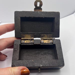 May include: Small, open wooden box with a hinged lid and brass hardware. The box is dark brown with a rectangular shape. The lid is open, revealing the interior. The box has a small brass clasp and hinges.