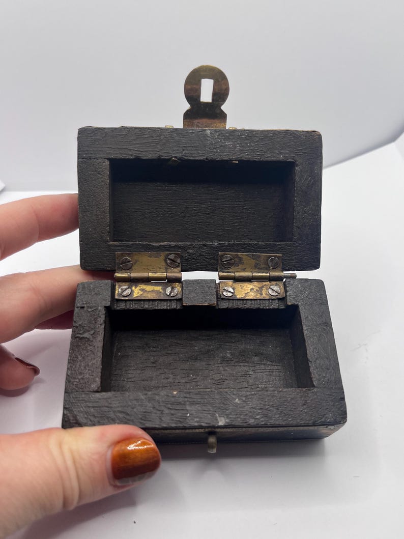 May include: Small, open wooden box with a hinged lid and brass hardware. The box is dark brown with a rectangular shape. The lid is open, revealing the interior. The box has a small brass clasp and hinges.