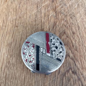 May include: A round silver-toned compact mirror featuring a decorative surface with alternating sections of engraved floral scrollwork, diagonal line patterns, and solid red and black enamel accents arranged in a geometric, segmented design.