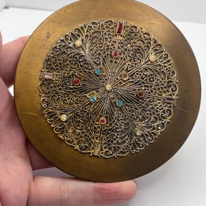 May include: A round, antique compact mirror with a brass-toned finish. The center features an ornate, filigree design with colorful rhinestones in red, blue, and pink. The compact is held in a hand.