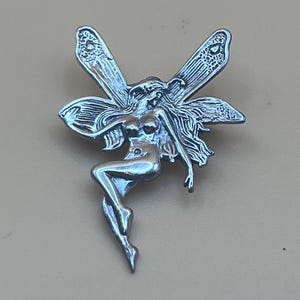 May include: A silver-toned fairy pin with detailed wings and flowing hair. The fairy is depicted in a seated pose, with one leg extended. The pin has a polished finish and is set against a neutral background.