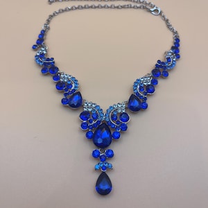 May include: A silver-toned necklace with a cascade of blue gemstones. The necklace features a series of smaller blue stones leading to larger, teardrop-shaped gems at the center. The design is symmetrical and elegant, suitable for formal wear.