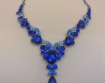 May include: A silver-toned necklace with a cascade of blue gemstones. The necklace features a series of smaller blue stones leading to larger, teardrop-shaped gems at the center. The design is symmetrical and elegant, suitable for formal wear.