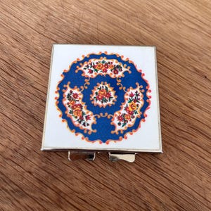 May include: A square, silver-framed compact mirror with a white top featuring a blue floral design. The design includes orange, yellow, and red flowers arranged in a circular pattern. The compact is open, revealing the mirror.