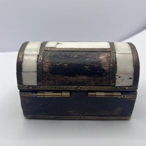 May include: A small, decorative wooden chest with a hinged lid. The chest features a dark wood base and a curved top with alternating sections of white and dark wood. Metal bands and hinges add detail.