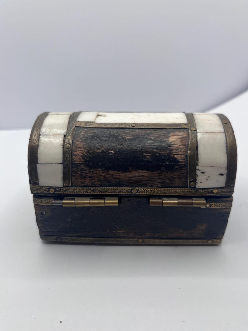 May include: A small, decorative wooden chest with a hinged lid. The chest features a dark wood base and a curved top with alternating sections of white and dark wood. Metal bands and hinges add detail.