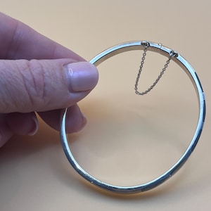 May include: A silver-colored bangle bracelet with a safety chain. The bracelet is round and has a simple, elegant design. The background is a solid, light orange color.