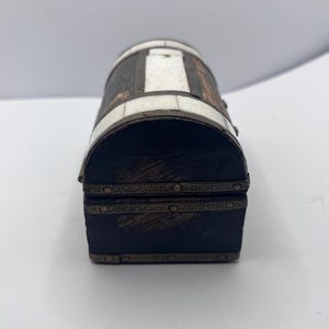 May include: A small, decorative chest with a barrel-shaped top. The chest is primarily dark brown with white rectangular inlays and bronze-colored metal trim. The front and back are dark brown with bronze-colored trim.