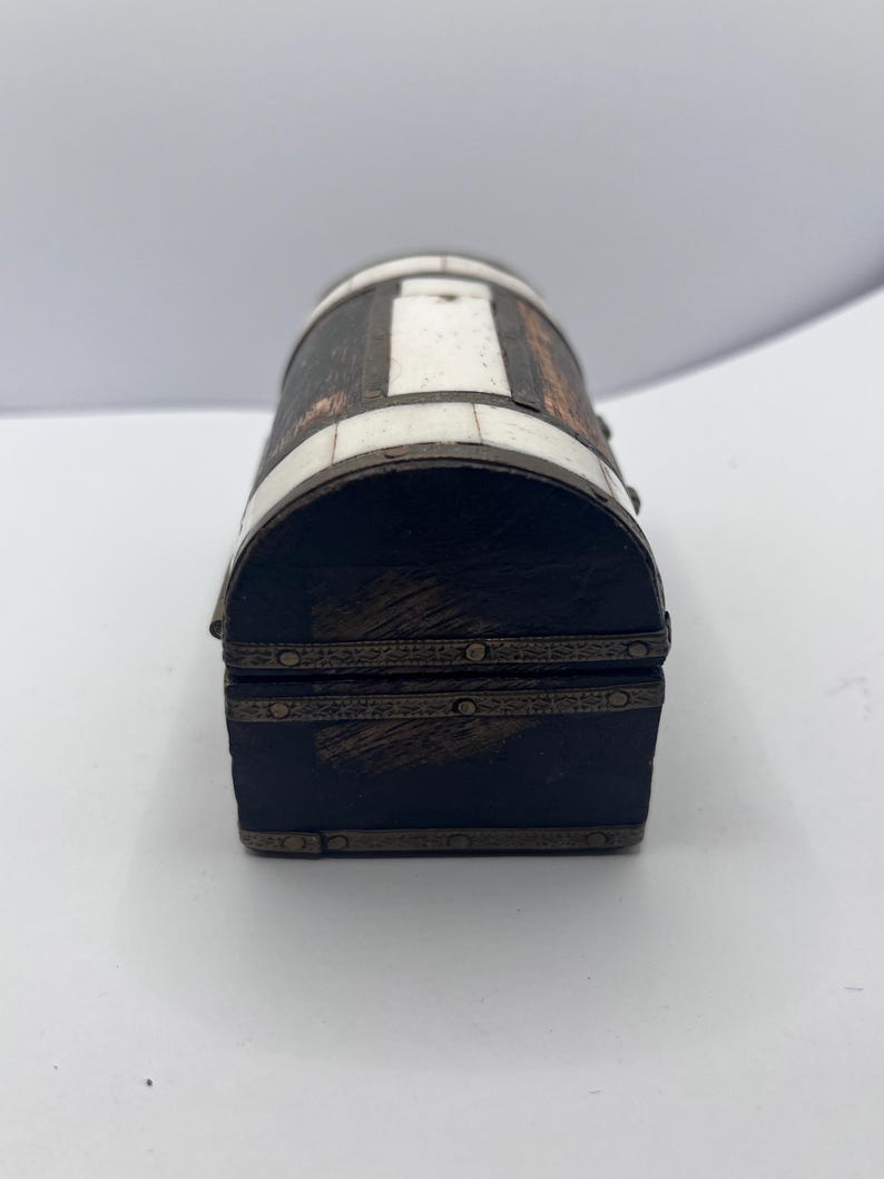 May include: A small, decorative chest with a barrel-shaped top. The chest is primarily dark brown with white rectangular inlays and bronze-colored metal trim. The front and back are dark brown with bronze-colored trim.