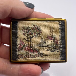May include: A small, rectangular, vintage compact with a gold-tone metal frame. The center features a tapestry scene with trees, buildings, and red and beige hues. The edges are black. The compact is held by a hand.