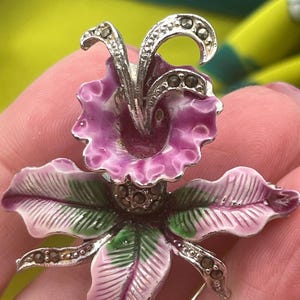 May include: A vintage floral brooch featuring a detailed orchid design. The brooch has a silver-tone metal base with a purple and pink enamel flower and green leaves. Accents include small, dark-colored stones.