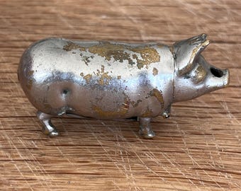 May include: A small, metallic pig figurine featuring a silver-toned finish with visible patches of worn gold-colored metal throughout its body. The pig is depicted in a standing profile position, showing its snout, ears, and four legs in detail.