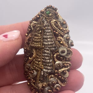 May include: An ornate, vintage brooch with a detailed, textured design. The brooch features a central tree-like motif in gold and silver threads, surrounded by gold and bronze beads and sequins. A small green gem is set at the top.