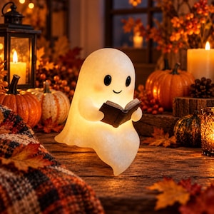 May include: A glowing, translucent white ghost figurine with a smooth, rounded head and small black eyes, holding a small, open dark-colored book. The ghost emits a warm, soft light from within, creating a gentle illumination.