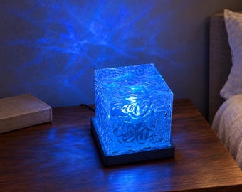 Op de afbeelding: Een vierkante glazen lamp met een getextureerd, golvend oppervlak dat een felblauw licht uitstraalt. De lamp staat op een donkere voet en projecteert een blauw, waterachtig patroon op de muur. De tekst Olive & Loom Finds is zichtbaar.