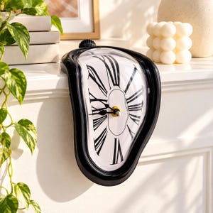 May include: A black melting-style analog clock with a white face and black Roman numerals. The clock features a curved, distorted frame designed to sit on the edge of a flat surface, with black hour and minute hands and a gold center.