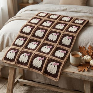 Könnte beinhalten: Eine Häkeldecke aus quadratischen Motiven mit weißen Geisterfiguren auf dunkelbraunem Hintergrund. Jeder Geist trägt eine kleine rosa Schleife auf dem Kopf. Die Decke ist mit einem hellbraunen Rand zwischen den Quadraten versehen.
