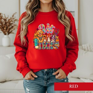 Puede incluir: Una sudadera roja de manga larga con un estampado gráfico colorido con el texto Vet Tech. El diseño incluye ilustraciones de un golden retriever, un conejo y un bulldog francés rodeados de calabazas, libros y un estetoscopio.
