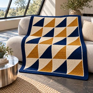 Puede incluir: Una manta de ganchillo rectangular con un patrón geométrico de triángulos azul marino, amarillo mostaza y color crema dispuestos en cuadrícula. La manta está rematada con un borde azul marino sólido que enmarca todo el diseño.
