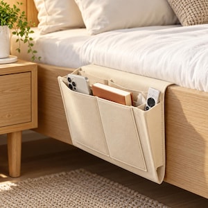 May include: A beige felt bedside storage organizer hanging from the side of a wooden bed frame. The organizer features multiple compartments holding a smartphone, a small book, and a remote control, providing accessible storage space.