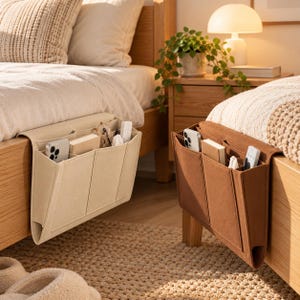 Può includere: Due organizer da comodino in feltro, uno beige e uno marrone, appesi al lato di telai di letto in legno. Ogni organizer presenta diverse tasche contenenti uno smartphone, un taccuino, occhiali e un telecomando.