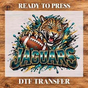 May include: A DTF transfer sheet featuring a snarling jaguar with spotted fur holding a brown football. The design includes teal and gold paint splashes and the word JAGUARS in bold, outlined lettering. Text reads READY TO PRESS.
