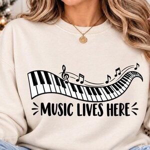 May include: A cream-colored crewneck sweatshirt featuring a black graphic of a wavy piano keyboard with musical notes floating above it. Below the keyboard, the text reads "MUSIC LIVES HERE" in a bold, black, sans-serif font.