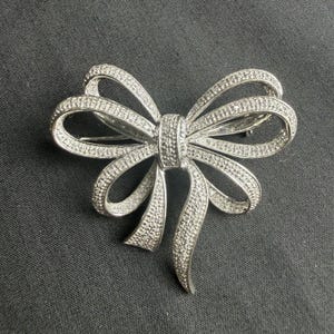 May include: A silver-toned metal brooch shaped like a bow, featuring a textured surface covered in numerous small, clear, sparkling stones. The design includes two main loops and two trailing ends, all encrusted with the reflective stones.