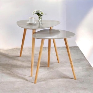 May include: A set of two nesting tables with smooth, light gray, triangular-shaped tops and three tapered wooden legs each. A white ceramic cup and a small glass vase with white flowers sit on the larger table surface.