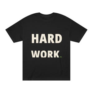 Può includere: Una t-shirt nera a maniche corte con il testo HARD WORK in grandi lettere maiuscole color bianco sporco centrate sul petto. Un piccolo cerchio verde oliva pieno è posizionato immediatamente a destra della parola WORK.