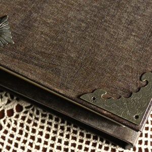 May include: A brown leather-bound journal with metal corner accents and a decorative metal piece on the front cover. The journal is resting on a white crocheted doily.